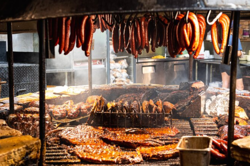 A smoky barbecue grill filled with various meats, including sausages and ribs, hanging above and sizzling on the grill.