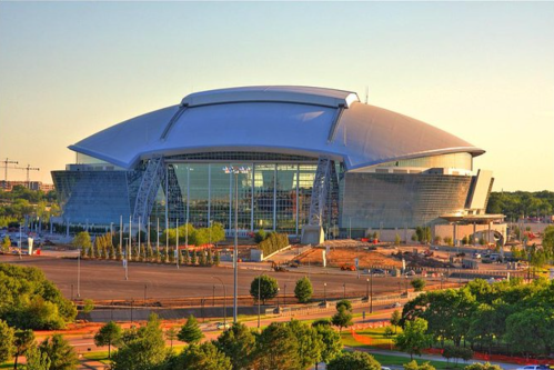 Aerial view of a modern stadium with a large, curved roof, surrounded by trees and a clear sky.