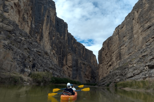 A person kayaks through a narrow canyon with steep rock walls and a cloudy sky above.