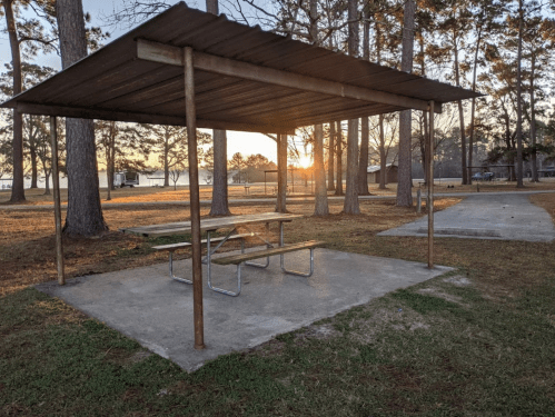 A picnic area with a shelter and table, surrounded by trees at sunset near a lake.