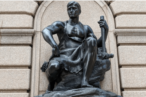 A bronze statue of a muscular figure seated on a rock, holding a tool, symbolizing strength and craftsmanship.