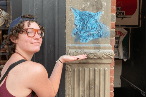 A person with curly hair and glasses poses next to a blue chalk drawing of a sleeping cat on a wall.