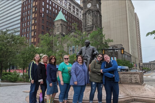 A group of eight women poses together near a statue in an urban setting, surrounded by trees and buildings.