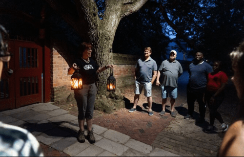 A tour guide holds lanterns while leading a group of people at night near a tree and brick wall.