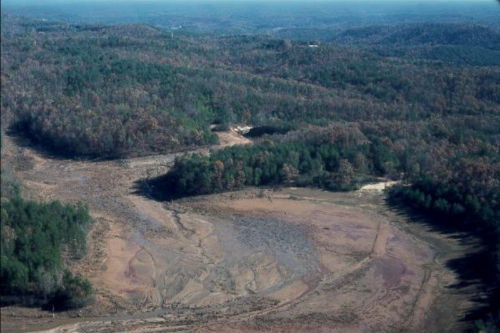 Aerial view of a dry riverbed surrounded by lush green forests and rolling hills in the background.
