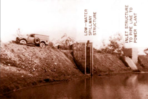Black and white photo of a low-water spillway structure with a vintage truck beside a river.
