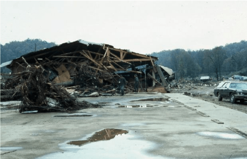 A collapsed building surrounded by debris and muddy ground, with a car parked nearby and trees in the background.