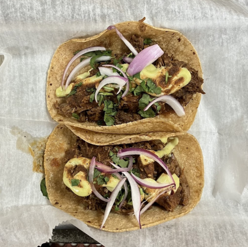 Two tacos filled with shredded meat, topped with onions, cilantro, and slices of avocado, served on a paper-lined tray.
