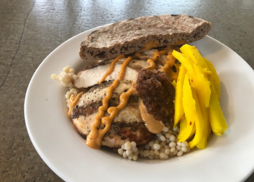 Grilled chicken with peanut sauce, couscous, mango slices, and flatbread on a white plate.
