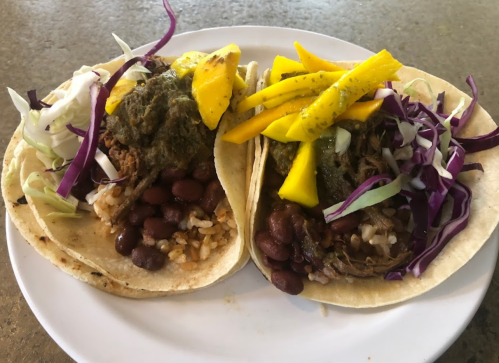Two tacos on a plate, filled with rice, beans, shredded meat, green sauce, and topped with mango and cabbage.