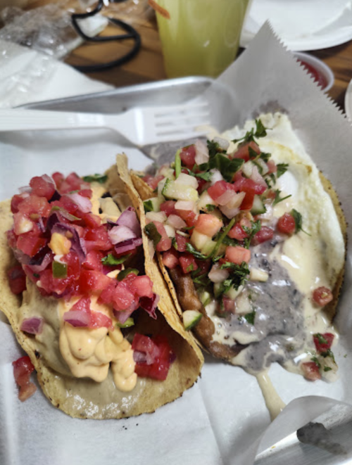 Two tacos on a white plate, one topped with creamy sauce and salsa, the other with black beans and fresh pico de gallo.