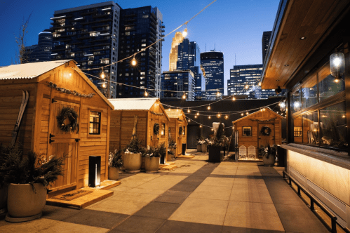 Cozy wooden cabins with festive decor on a rooftop, city skyline illuminated at dusk in the background.
