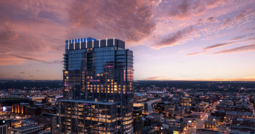 A modern skyscraper illuminated at dusk, surrounded by city lights and a colorful sunset sky.