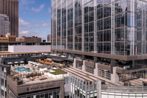 A modern hotel with a pool and outdoor seating, surrounded by glass buildings and a clear blue sky.