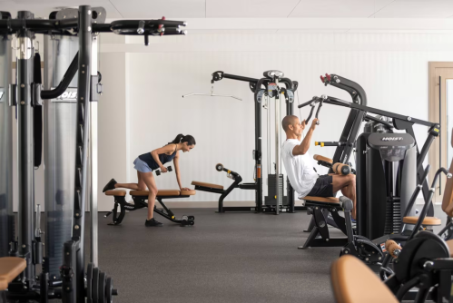 Two people exercising in a modern gym, one using a weight machine and the other performing a bent-over row.