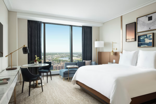 A modern hotel room featuring a king bed, seating area, desk, and large window with a city view.