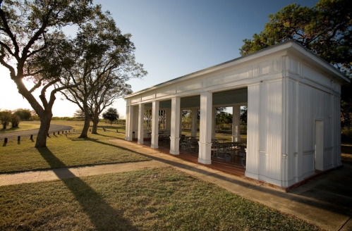 A white pavilion with columns stands in a grassy area, surrounded by trees and a clear blue sky.