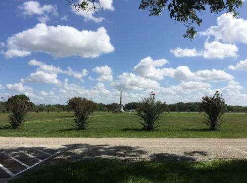 A grassy field with a tall monument in the distance, surrounded by trees and fluffy clouds in a blue sky.