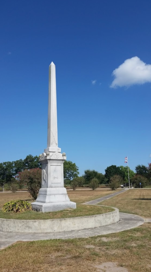 A tall white obelisk monument stands in a grassy area under a clear blue sky with a few clouds.