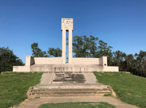 A tall monument with a decorative top, set on a grassy area with steps leading up to it under a clear blue sky.