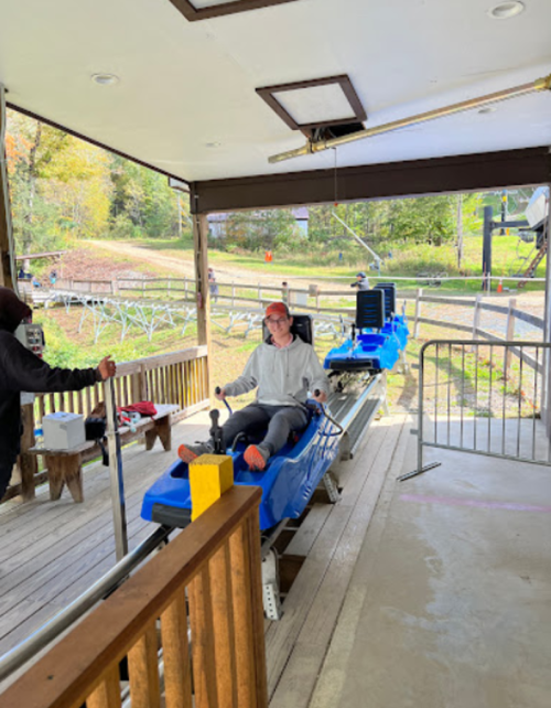 A person sits in a blue sled at a loading area for a mountain coaster, with a staff member nearby.
