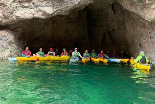 A group of kayakers in colorful kayaks gathered in a cave with clear green water.