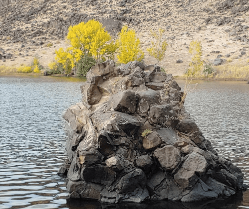 A rocky outcrop rises from a calm lake, surrounded by autumn-colored trees and a dry, hilly landscape.