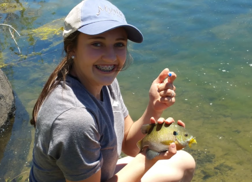 A smiling girl with braces holds a fish by a lake, wearing a cap and sitting on the shore.