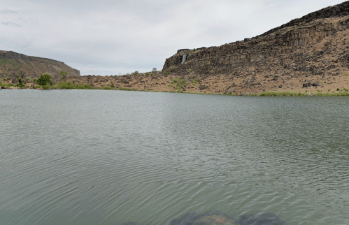A calm lake surrounded by rocky cliffs and sparse vegetation under a cloudy sky.