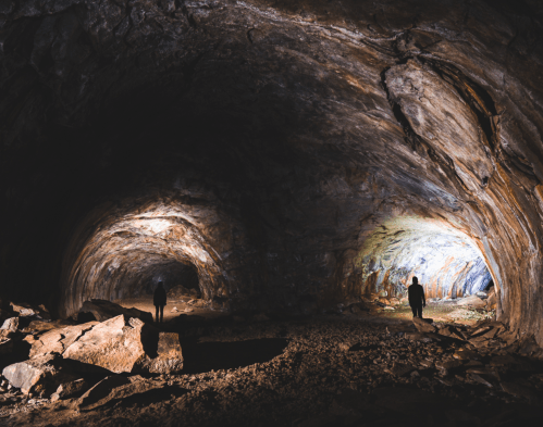 Two silhouetted figures stand in a dimly lit cave, surrounded by rocky formations and illuminated walls.