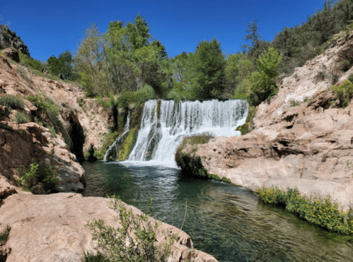 A serene waterfall cascades into a clear pool, surrounded by lush greenery and rocky terrain under a bright blue sky.