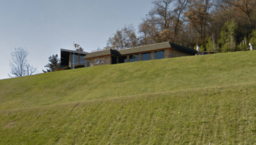 A modern building on a grassy hill, surrounded by trees, with a flagpole in front and a car parked nearby.