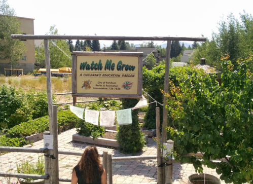 A sign reading "Watch Me Grow" at a children's education garden, surrounded by greenery and pathways.