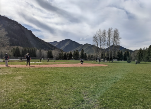 A baseball field with players in action, surrounded by mountains and trees under a cloudy sky.