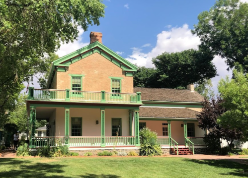 Historic two-story house with green trim, surrounded by trees and a grassy lawn under a partly cloudy sky.