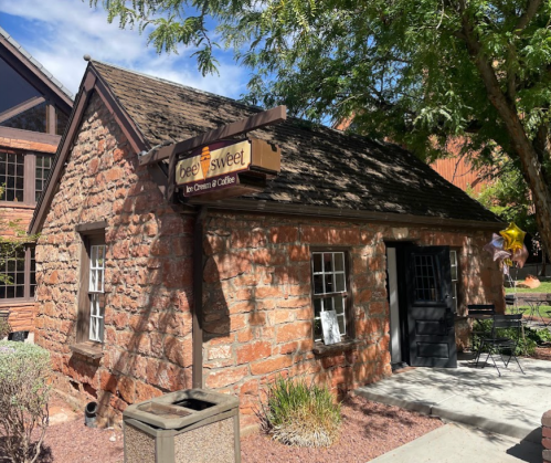 A small stone building with a sign reading "Bee Sweet" and outdoor seating, surrounded by greenery.