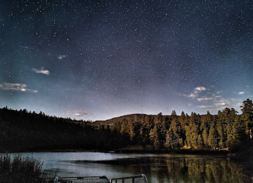 A serene lake surrounded by trees under a starry night sky, with mountains in the background.