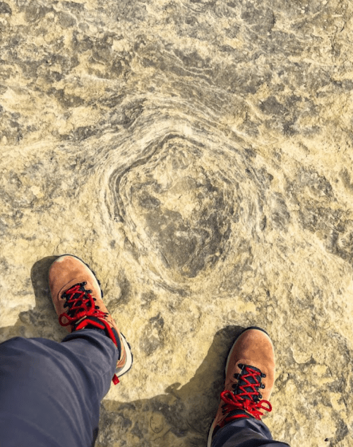 A person's feet in hiking boots stand on a rocky surface with a circular pattern.