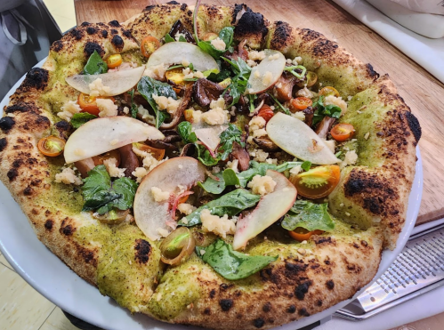 A freshly baked pizza topped with greens, sliced apples, cherry tomatoes, and crumbled cheese on a charred crust.