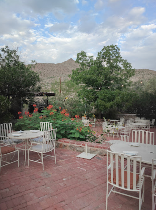 Outdoor dining area with white chairs and tables, surrounded by greenery and mountains under a cloudy sky.