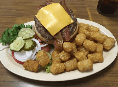 A cheeseburger with bacon, lettuce, tomato, and pickles, served with a side of crispy tater tots on a plate.