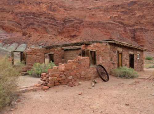 Abandoned stone building with a partially collapsed wall, set against red rock formations and sparse vegetation.