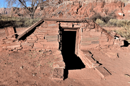 A small, weathered stone structure with a wooden door, set against a backdrop of red rock formations and sparse vegetation.