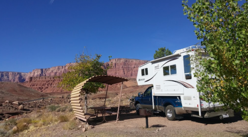 A camper parked under a wooden shelter with a scenic red rock backdrop and clear blue sky.