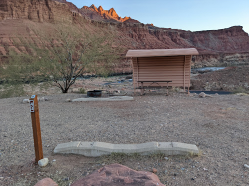 A campsite with a shelter, picnic table, and fire pit, surrounded by rocky terrain and mountains at sunset.