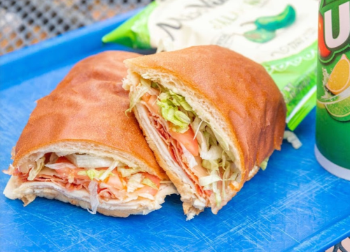 A close-up of a sandwich filled with lettuce, tomato, and sliced meat, alongside a bag of chips and a drink.