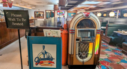 A retro diner entrance with a "Please Wait to Be Seated" sign and a vintage jukebox nearby. Colorful carpet visible.