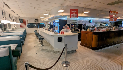 A retro diner interior featuring a long counter, teal booths, and vintage decor, with an American flag in the background.