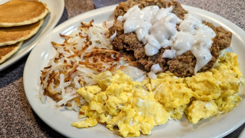 A plate of breakfast featuring fried chicken with gravy, scrambled eggs, hash browns, and pancakes on the side.