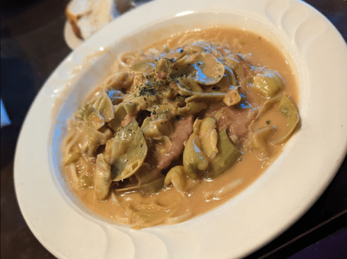 A plate of pasta with artichokes and a creamy sauce, garnished with herbs, served with a slice of bread in the background.